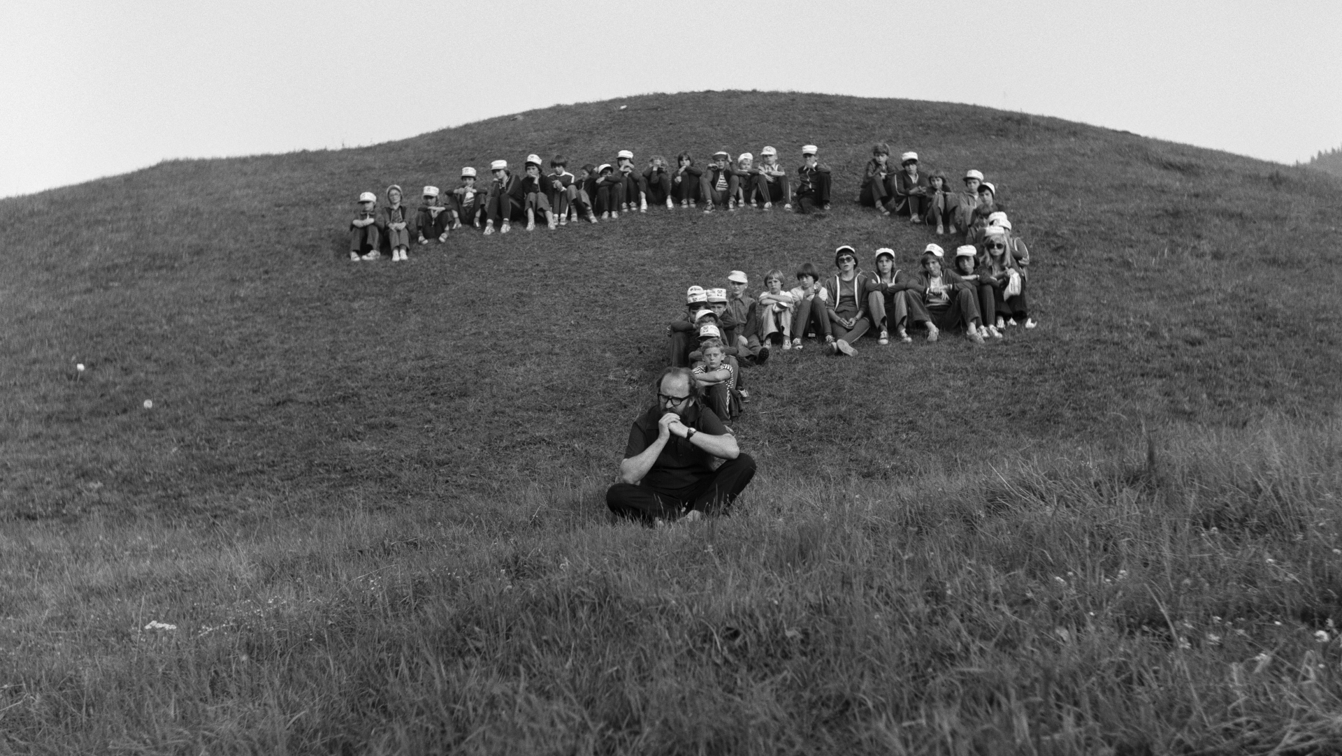 Czarno-biała fotografia przedstawiająca happening Juliusa Kollera. Na wzgórzu dziecięce postaci tworzą kształt wielkiego znaku zapytania. W miejscu kropki siedzi Juliusz Koller, autor happeningu. 