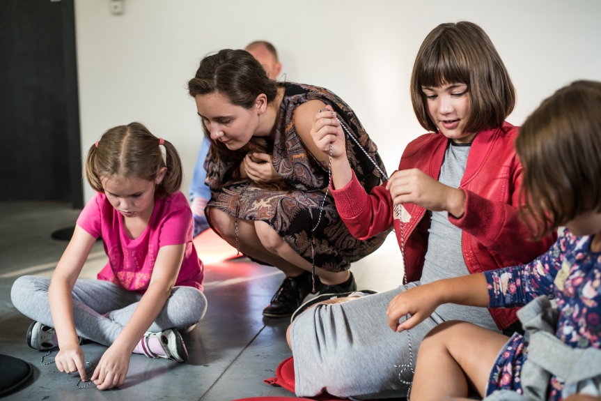 Zdjęcie. Grupa małych dzieci na wystawie, towarzyszy im edukatorka, która patrzy z zainteresowaniem jak jedno z nich rysuje na kartce. 