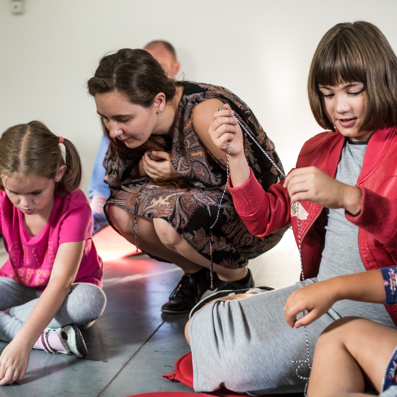 Zdjęcie. Grupa małych dzieci na wystawie, towarzyszy im edukatorka, która patrzy z zainteresowaniem jak jedno z nich rysuje na kartce. 