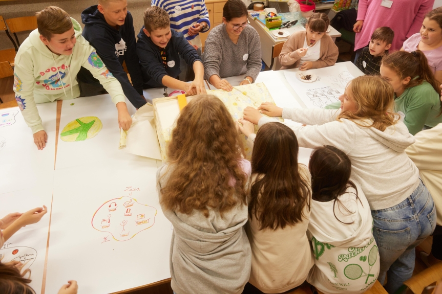 Zdjęcie. Grupa uczniów pochyla się nad wspólnym biurkiem, na którym leży pudełko, które odpakowują. 