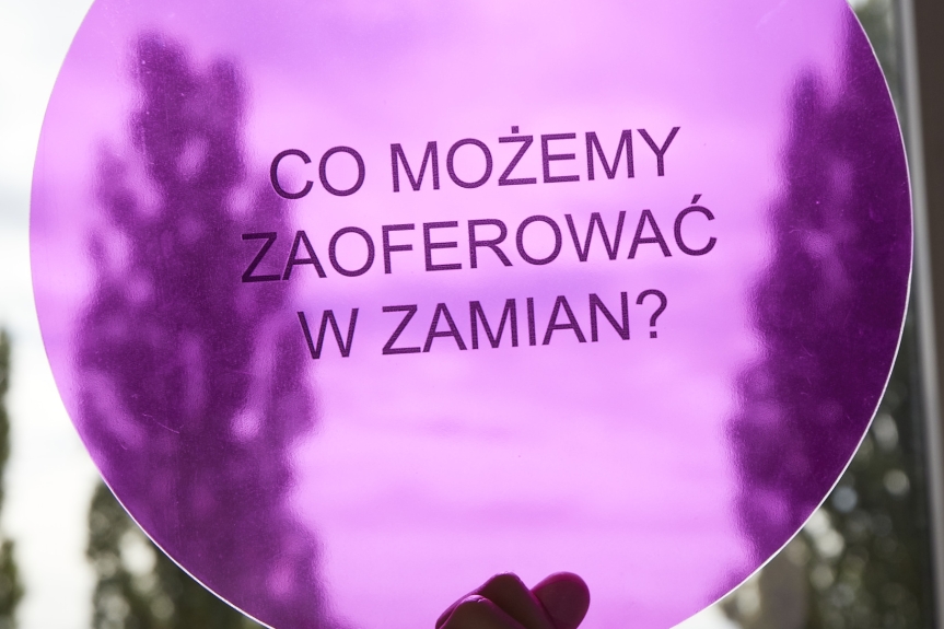 Zdjęcie. Różowe, półprzezroczyste kółko trzymane w dłoni, na tle okna, kole napis Co możemy zaoferować w zamian. 