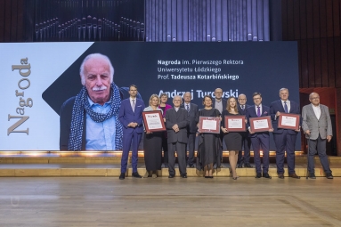 kilkanaście osób na scenie, niektóre z nich trzymają dyplomy, w tle ekran, na którym wyświetla się twarz mężczyzny i napis Nagroda